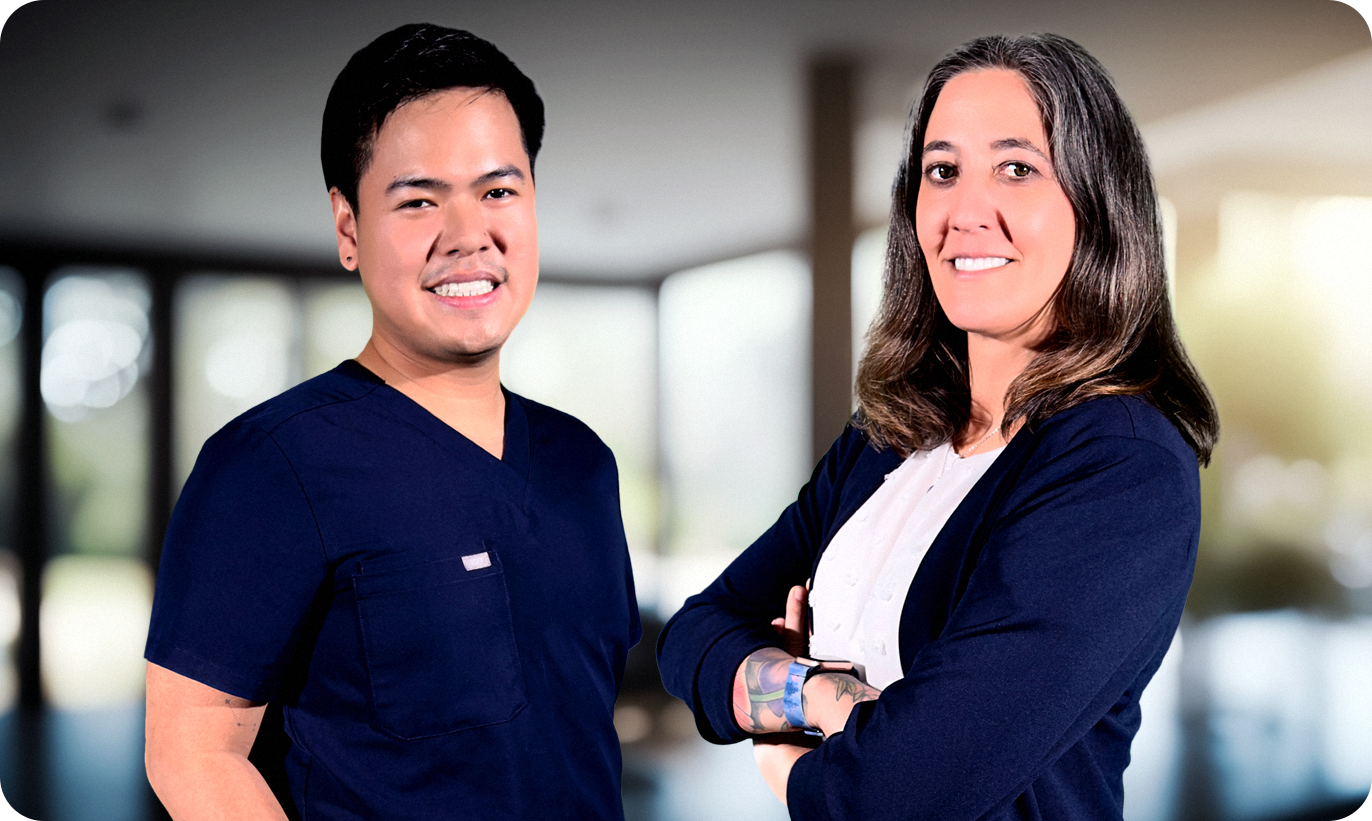 Two professionally dressed individuals pose in a modern, well-lit indoor setting. The man on the left wears navy blue medical scrubs and smiles warmly. The woman on the right wears a white blouse with a navy blue blazer, has a tattoo on her forearm, and crosses her arms while smiling confidently. The blurred background features large windows with natural light.