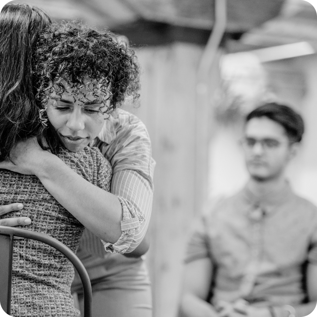 A black and white photo of a curly-haired person embracing someone tightly with an emotional expression, eyes closed and brows furrowed. In the background, a young man wearing glasses and a button-up shirt sits, observing the moment. The setting appears to be indoors with a rustic ambiance.